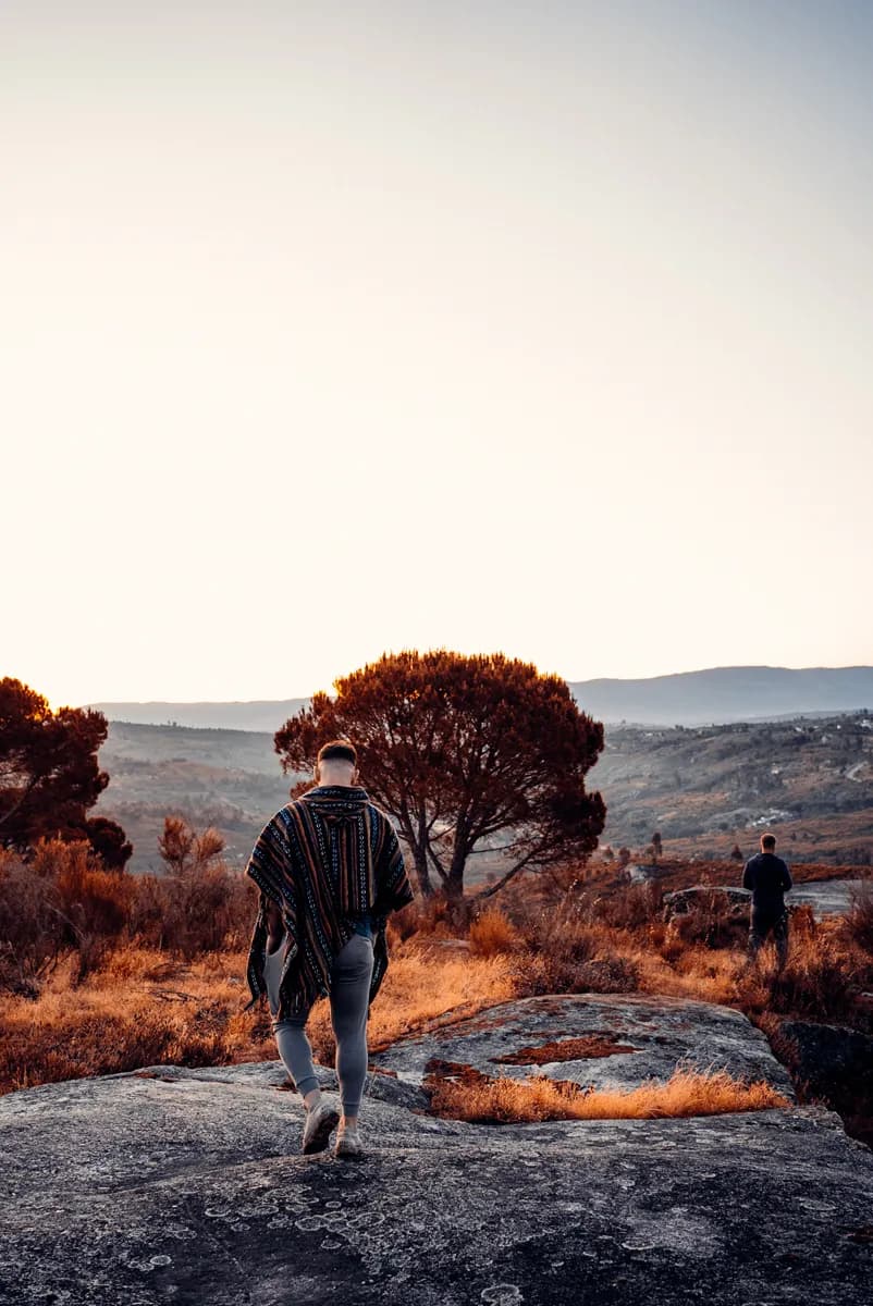 Guest in a poncho walking across rocky hilltop toward a panoramic valley view at golden hour, Central Portugal